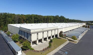 Alt text Large modern food-grade distribution warehouse with multiple truck bays and semi-trailers lined up for loading in a wooded industrial area.