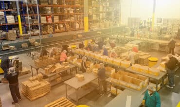 Busy integrated supply chain warehouse floor with workers packaging products at tables near a conveyor belt and high shelves.