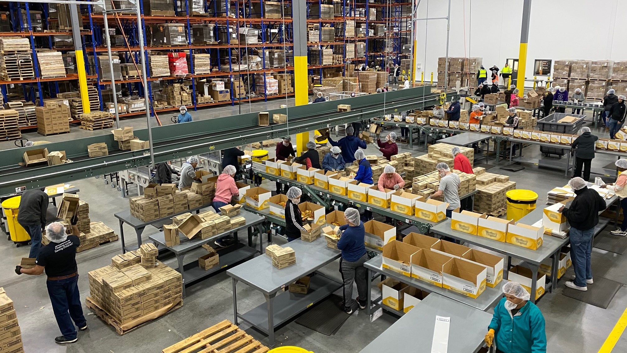 Team members pack cartons along conveyors on a co-packing floor run by a 3PL provider, with tall racking behind them